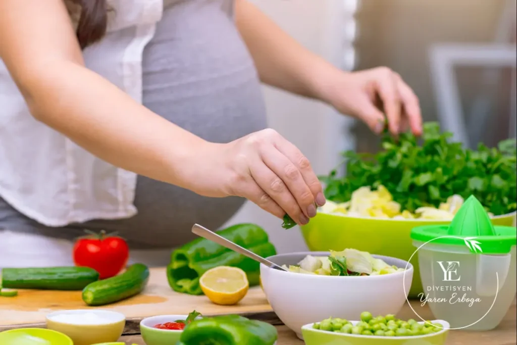 Hamile kadın, salata kasesine yeşillik eklerken; tezgâhta domates, salatalık, limon ve sebzeler