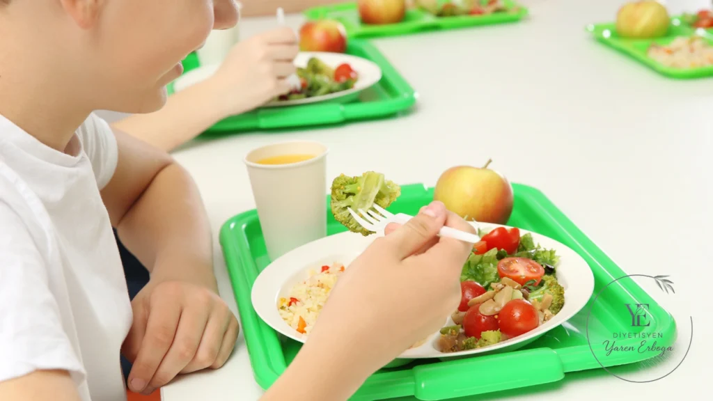 Çocuk, okul yemekhanesinde pilav, brokoli ve salata bulunan öğününden çatalıyla yemek yiyor