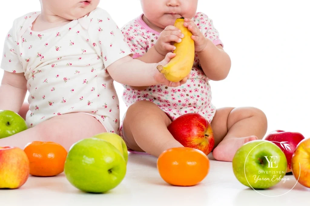 İki bebek, çevrelerine dizilmiş elma, muz ve portakal gibi meyvelerin yanında oturuyor