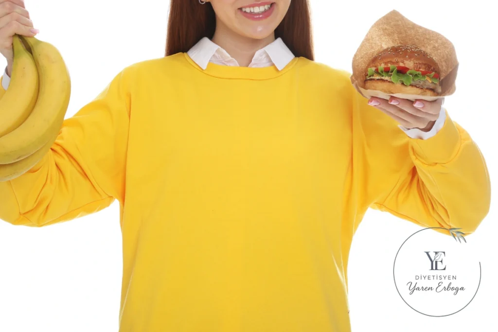 Sarı kazaklı bir kişi, bir elinde muz diğer elinde sebzeli hamburger tutuyor