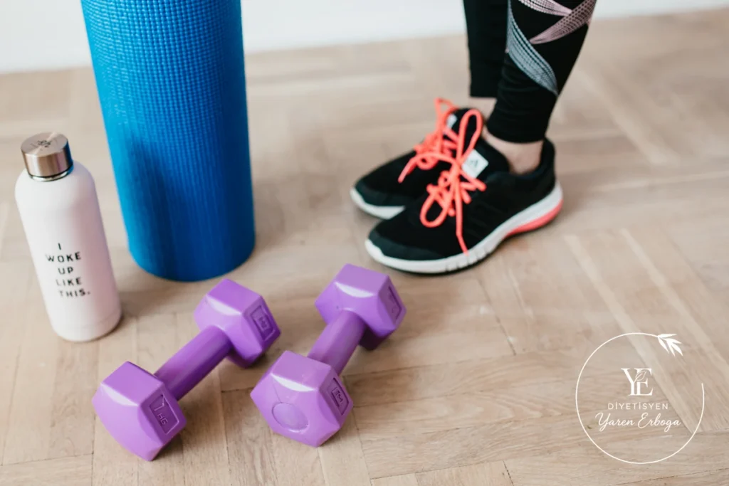 Egzersiz için hazırlanmış mor dambıllar, su matarası ve yoga matının yanında spor ayakkabılı kişi