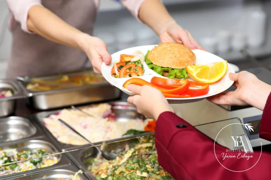 Yemekhanede bir kişiye hamburger, salata, biber ve portakal dilimi bulunan tabak servis ediliyor